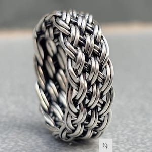 以下が含まれることがあります： 編み込みデザインのシルバーカラーのリング。リングは、絡み合ったストランドの詳細なパターンを備え、テクスチャのある表面を作り出しています。リングはバンドスタイルで、太い編み込みの外観です。