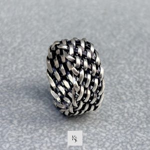 以下が含まれることがあります： 編み込みデザインのシルバーカラーのリング。リングは丸みを帯びた形状で、金属ストリップを織り交ぜて作られており、テクスチャード加工された表面を作り出しています。リングは灰色の表面に表示されています。