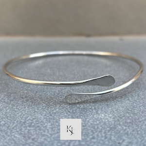 Op de afbeelding: Een zilverkleurige open manchetarmband. De armband heeft een eenvoudig, elegant ontwerp met afgeplatte, druppelvormige uiteinden. De sieraden worden gepresenteerd op een grijze ondergrond.
