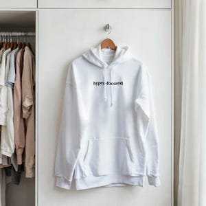 Op de afbeelding: Een witte hoodie met een voorzak en een capuchon met trekkoord. De zwarte tekst "hyper-focused" is op de borst gedrukt. De hoodie hangt aan een houten hanger.