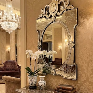 May include: An elaborate antique mirror with an oval centre and etched detailing is mounted on a gold-toned wall. A marble-topped table below holds white orchids in a patterned vase, a smaller vase, and stacked books.