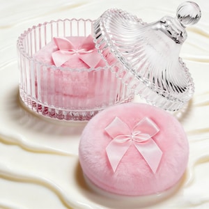 Può includere: Un barattolo di vetro trasparente con coperchio, contenente un piumino rosa con un fiocco di raso. Un altro piumino rosa con un fiocco è posizionato fuori dal barattolo. Il barattolo e il piumino sono su una superficie bianca.