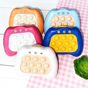 Op de afbeelding: Vijf kleurrijke draagbare sensorische speeltjes in oranje, blauw, roze, wit en blauw met geel. Elk speeltje heeft een raster van cirkelvormige bellen en bedieningsknoppen. De speeltjes zijn gerangschikt op een roze en witte geruite ondergrond.