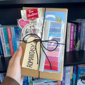 May include: A gift box tied with black string, with a tag reading "Romantasy." Inside are a bookmark, markers, and a Tim Hortons packet. Bookshelves filled with books are in the background.