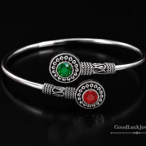 Może przedstawiać: Srebrna bransoletka typu bangle z dwoma okrągłymi elementami dekoracyjnymi. Jeden element ma zielony kamień szlachetny, drugi czerwony. Bransoletka ma otwartą konstrukcję i jest umieszczona na czarnym tle.