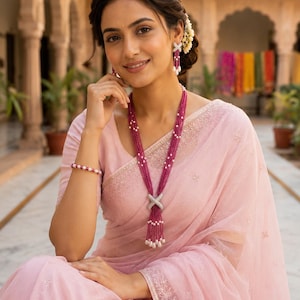 May include: A light pink saree with intricate embroidery paired with a long multi-strand necklace featuring deep pink beads, small white pearls, and a central silver-toned cross-shaped pendant. Matching drop earrings and a beaded bracelet complete the set.
