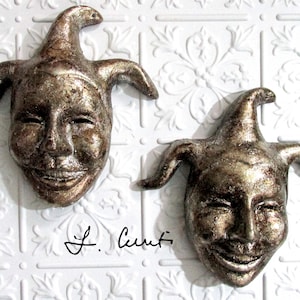 May include: Two gold-painted ceramic jester masks with a whimsical expression. The masks have exaggerated features and are mounted on a white background.