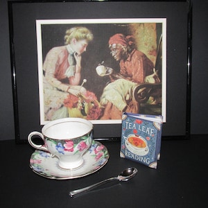 May include: A framed vintage illustration of two women, one in a red dress and the other in a pink dress, sitting together with a cup of tea. A small blue book with the title "Tea Leaf Reading" is in the foreground.