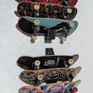 Peut inclure: Une collection de mini-skateboards de différents motifs et couleurs, montés sur un mur. Les planches présentent des graphiques, des logos et du texte, dont "LUTHERAN" et "ELEMENT". Les roues sont blanches et jaunes. Les planches sont maintenues par des supports noirs.