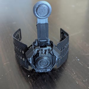 May include: A black, 3D-printed wrist-mounted device with a central circular component and a raised, textured top. The device has a complex design with multiple articulated parts and a textured wristband. The overall aesthetic suggests a futuristic or technological theme.
