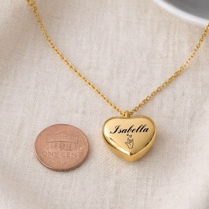 May include: A gold-colored heart-shaped pendant necklace with the name "Isabella" engraved on it, along with a hand symbol and a heart. A penny is next to the pendant. The necklace is on a white surface.