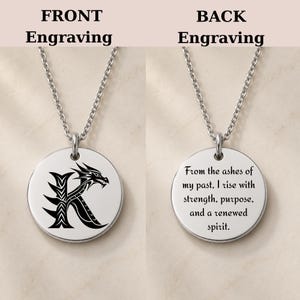 May include: A silver-toned circular pendant necklace featuring a black dragon-shaped letter K on the front and the engraved text: From the ashes of my past, I rise with strength, purpose, and a renewed spirit on the back side of the metal disc.
