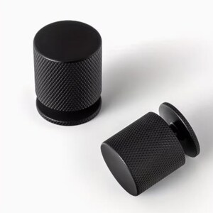 Puede incluir: Dos tiradores de armario negros con forma cilíndrica y superficie texturizada con patrón de diamantes. Uno está de pie, el otro tumbado. Los tiradores tienen una parte superior redondeada y una base más ancha.