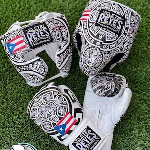 Könnte beinhalten: Ein Set aus weißem Boxkopfschutz und einem Paar Boxhandschuhen mit komplexen schwarz-weißen Mustern und der Flagge von Puerto Rico. Jedes Teil zeigt das Cleto Reyes Profesional Logo in schwarzer und weißer Schrift auf einem rechteckigen Aufnäher.