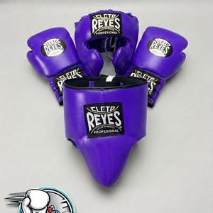 Peut inclure: Un ensemble d'équipement de boxe violet comprenant deux gants de boxe, un casque et une coquille de protection. Chaque pièce arbore l'écusson noir et blanc Cleto Reyes Profesional. Les articles sont disposés ensemble sur une surface neutre.