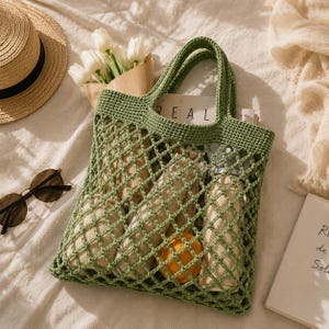 Styled flat lay of handmade crochet mesh tote bag with sunglasses, book, dried flowers, and neutral fabric background in warm natural lighting.