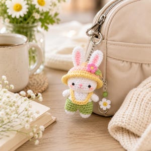 May include: A crochet bunny keychain featuring a white rabbit wearing a yellow sun hat with a small pink flower, green overalls, and a silver metal chain with a small white daisy charm attached to a beige leather handbag.