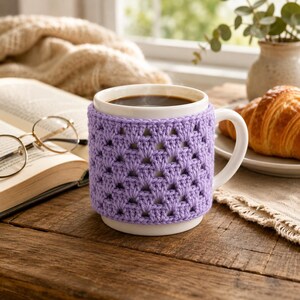 Può includere: Una tazza in ceramica bianca con un rivestimento all'uncinetto color viola chiaro con un motivo ripetitivo a piastrelle granny. La tazza ha un manico robusto ed è riempita con un liquido scuro, mostrando una fodera in filato testurizzata fatta a mano.