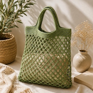 May include: A green crocheted tote bag with a diamond-patterned mesh design. The bag has two handles and is made of a soft, textured yarn. The bag is empty and sits on a white surface.