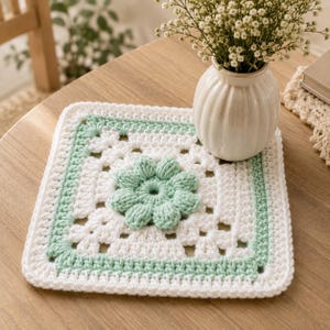 May include: A square crochet coaster featuring a white base with a central light green puff stitch flower motif. The border consists of alternating rows of white and light green yarn, creating a textured, geometric pattern with openwork details.