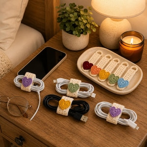 May include: A collection of cream-colored crochet cable organizers, each featuring a small, colorful heart accent in shades of purple, green, yellow, orange, and blue. These organizers are used to bundle white and black charging cables neatly.
