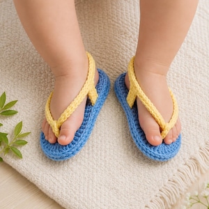 Könnte beinhalten: Ein Paar blaue und gelbe Baby-Häkelsandalen. Die Sandalen haben eine blaue Basis und gelbe Riemen. Die Sandalen befinden sich auf einer cremefarbenen Oberfläche. Die Sandalen sind als Flip-Flops gestaltet.