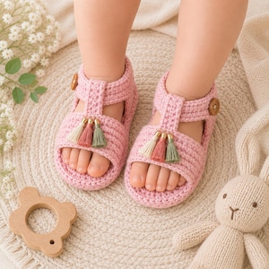 Puede incluir: Sandalias rosas de crochet para bebé, con diseño de tira en T. Cada sandalia presenta un cierre de botón de madera y tres pequeñas borlas en crema, rosa y verde. Las sandalias están diseñadas para un pie de bebé.