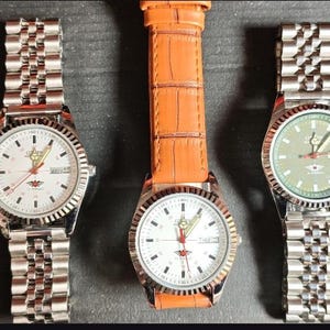 Pode incluir: Três relógios de pulso. Dois têm pulseiras de metal prateado e um tem uma pulseira de couro laranja. Os mostradores são brancos, com marcadores de hora pretos e ponteiros de segundos vermelhos. Os relógios são exibidos em uma superfície escura.