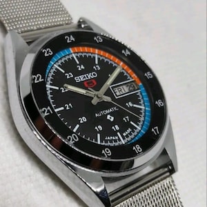 Pode incluir: Relógio automático Seiko prateado com mostrador preto. O mostrador apresenta algarismos brancos, um visor de dia e data e detalhes em azul e laranja. A pulseira é de malha.