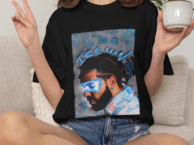 Puede incluir: Una camiseta negra con un estampado gr&aacute;fico de un hombre con barba, estilizado con acentos de aer&oacute;grafo azul sobre los ojos y el texto "ICEMAN" sobre la cabeza. El texto adicional incluye "MAY 15", "Papi" y "6GOD" en letras azules y rojas.