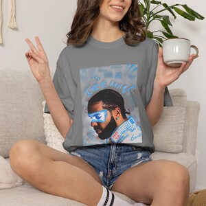 Puede incluir: Una camiseta gris carb&oacute;n de manga corta con un estampado gr&aacute;fico de un hombre con una m&aacute;scara pintada de azul sobre los ojos. El texto "ICE MAN" aparece en azul sobre el retrato, con texto adicional en azul como "MAY 15" y "6GOD" debajo.