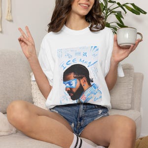 Puede incluir: Una camiseta blanca de manga corta con un estampado gr&aacute;fico de un hombre con una m&aacute;scara pintada de azul sobre los ojos. El texto "ICE MAN" est&aacute; impreso en azul sobre el retrato, rodeado de varios garabatos y notas azules a mano.