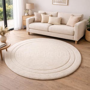 May include: A circular cream rug with a concentric design, placed on a wooden floor. The rug is in a living room with a beige sofa, cushions, and a side table with a lamp. The rug's diameter is approximately 200 cm.