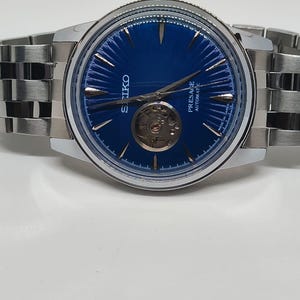 May include: A stainless steel Seiko Presage automatic watch with a blue dial. The watch features a visible escapement and a silver-tone bracelet. The dial has silver hands and markers. The watch is displayed on a white surface.