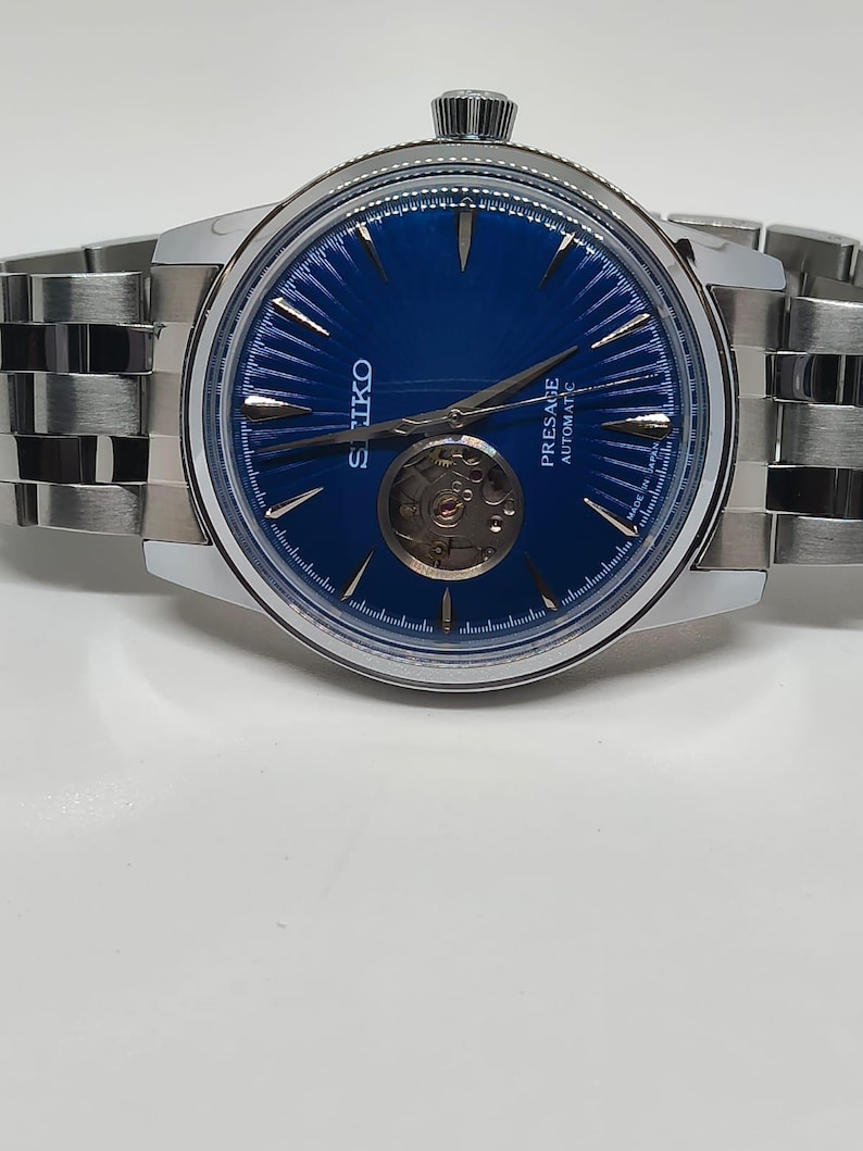 May include: A stainless steel Seiko Presage automatic watch with a blue dial. The watch features a visible escapement and a silver-tone bracelet. The dial has silver hands and markers. The watch is displayed on a white surface.
