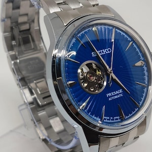 May include: A stainless steel Seiko Presage automatic watch with a vibrant blue dial. The watch features a visible escapement and a silver-toned link bracelet. The dial has a textured pattern and the words "Presage Automatic" are visible.