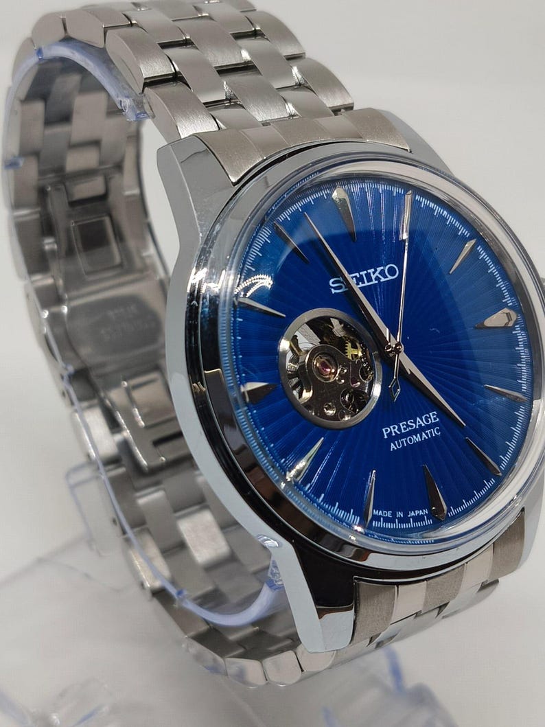 May include: A stainless steel Seiko Presage automatic watch with a vibrant blue dial. The watch features a visible escapement and a silver-toned link bracelet. The dial has a textured pattern and the words "Presage Automatic" are visible.