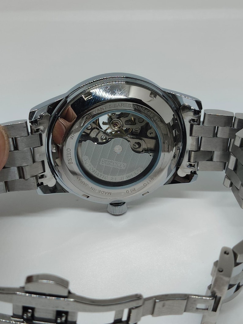 May include: A silver Seiko automatic wristwatch with a stainless steel strap. The watch has a transparent back, revealing the internal mechanism. The watch is marked "Made in Japan" and "Water Resistant 5 Bar".