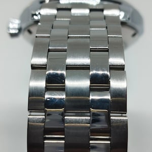 May include: Close-up of a stainless steel watch strap. The strap is made up of rectangular, interlocking links, reflecting light. The watch case is visible in the background. The watch strap is a classic design.