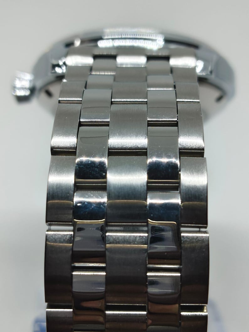 May include: Close-up of a stainless steel watch strap. The strap is made up of rectangular, interlocking links, reflecting light. The watch case is visible in the background. The watch strap is a classic design.