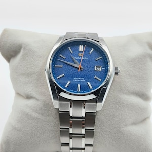 May include: A silver-tone stainless steel wristwatch with a textured blue dial, silver hour markers, and a date window at the three o'clock position. The dial features the text Grand Seiko, Automatic, and Hi-Beat 36000 with a thin orange second hand.