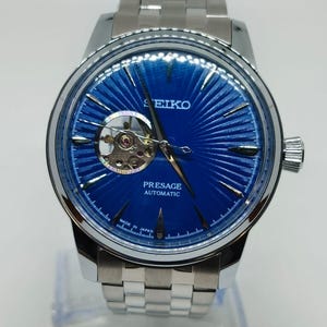 May include: A silver Seiko Presage automatic watch with a striking blue dial. The watch has a visible escapement and a stainless steel strap. The dial features a textured, sunburst pattern. The watch is marked "Made in Japan".