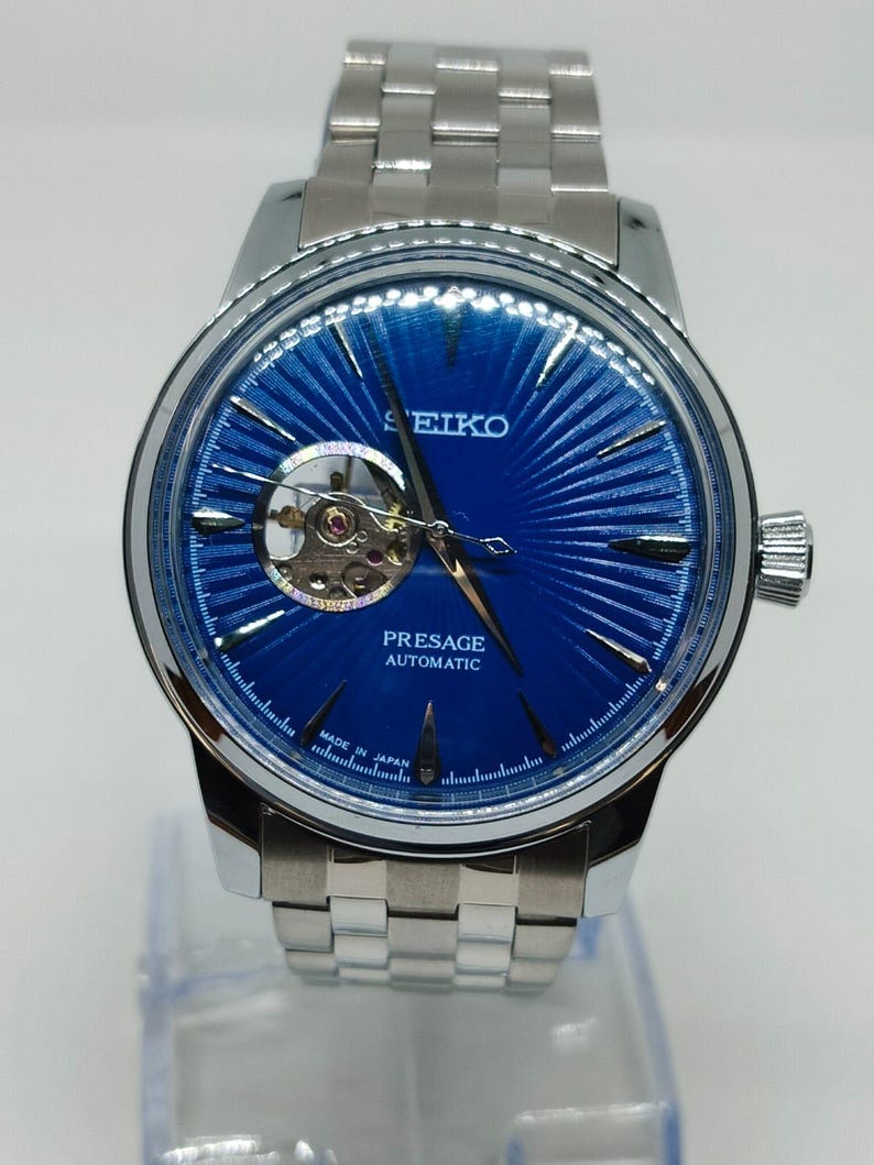 May include: A silver Seiko Presage automatic watch with a striking blue dial. The watch has a visible escapement and a stainless steel strap. The dial features a textured, sunburst pattern. The watch is marked "Made in Japan".