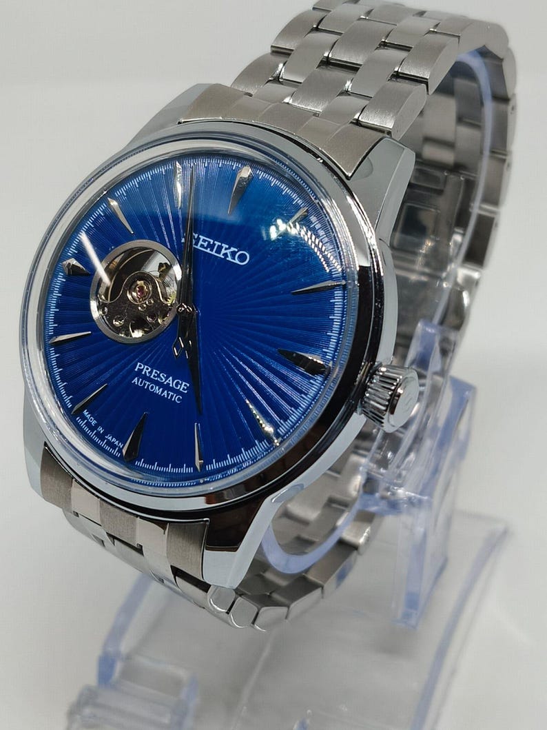 May include: A stainless steel Seiko Presage automatic watch with a striking blue dial. The watch has a visible escapement and a silver link bracelet. The dial has a textured pattern. The words "Presage Automatic" are visible.