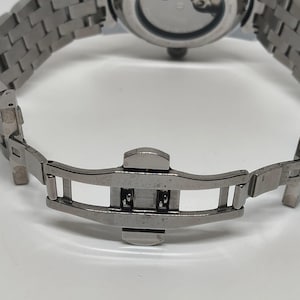 May include: Close-up of a silver stainless steel watch with a metal strap. The watch's clasp is open, revealing the intricate design of the strap and the watch's back.