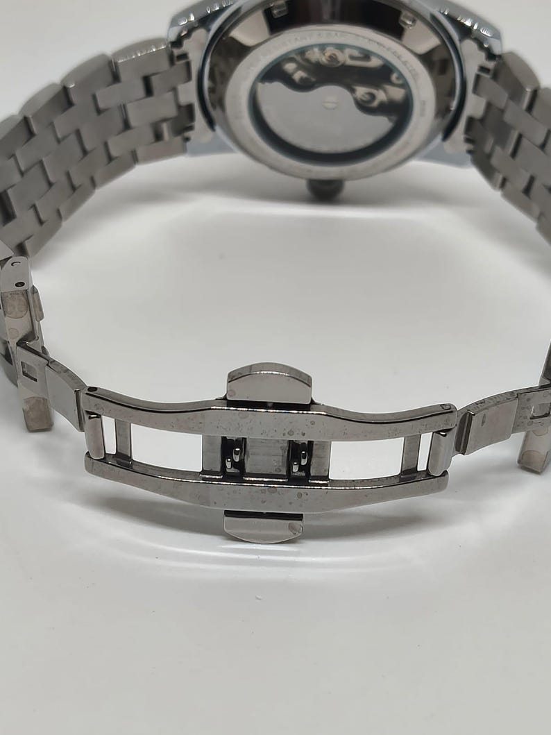May include: Close-up of a silver stainless steel watch with a metal strap. The watch's clasp is open, revealing the intricate design of the strap and the watch's back.