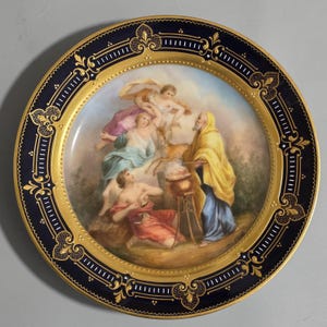 May include: A decorative porcelain plate with a dark blue border and gold accents. The center features a detailed painting of figures in classical attire, with angels and a man in a yellow robe. The plate has a glossy finish.
