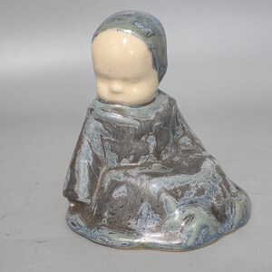 May include: A ceramic figurine of a seated figure wearing a hooded garment. The figure has a smooth, cream-coloured head and a textured, mottled glaze in shades of dark blue, grey, and brown covering the body and hood, creating a glossy finish.