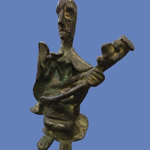 Puede incluir: Una escultura de bronce de una figura sentada con una cara larga y los brazos extendidos, sosteniendo un instrumento musical. La figura tiene una superficie texturizada y está montada sobre una base circular. La escultura tiene un aspecto envejecido.