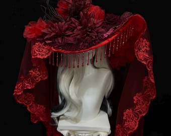 May include: A wide-brimmed red hat adorned with dark red fabric flowers and thin, dark twigs. The hat features a long, sheer red lace veil with scalloped edges and a fringe of small, dangling red beads hanging from the brim's inner edge.